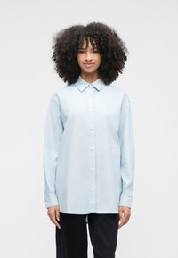 Kvinna med lockigt hår som bär en ljusblå skjorta med knappar och svarta byxor, stående mot en enkel vit bakgrund.