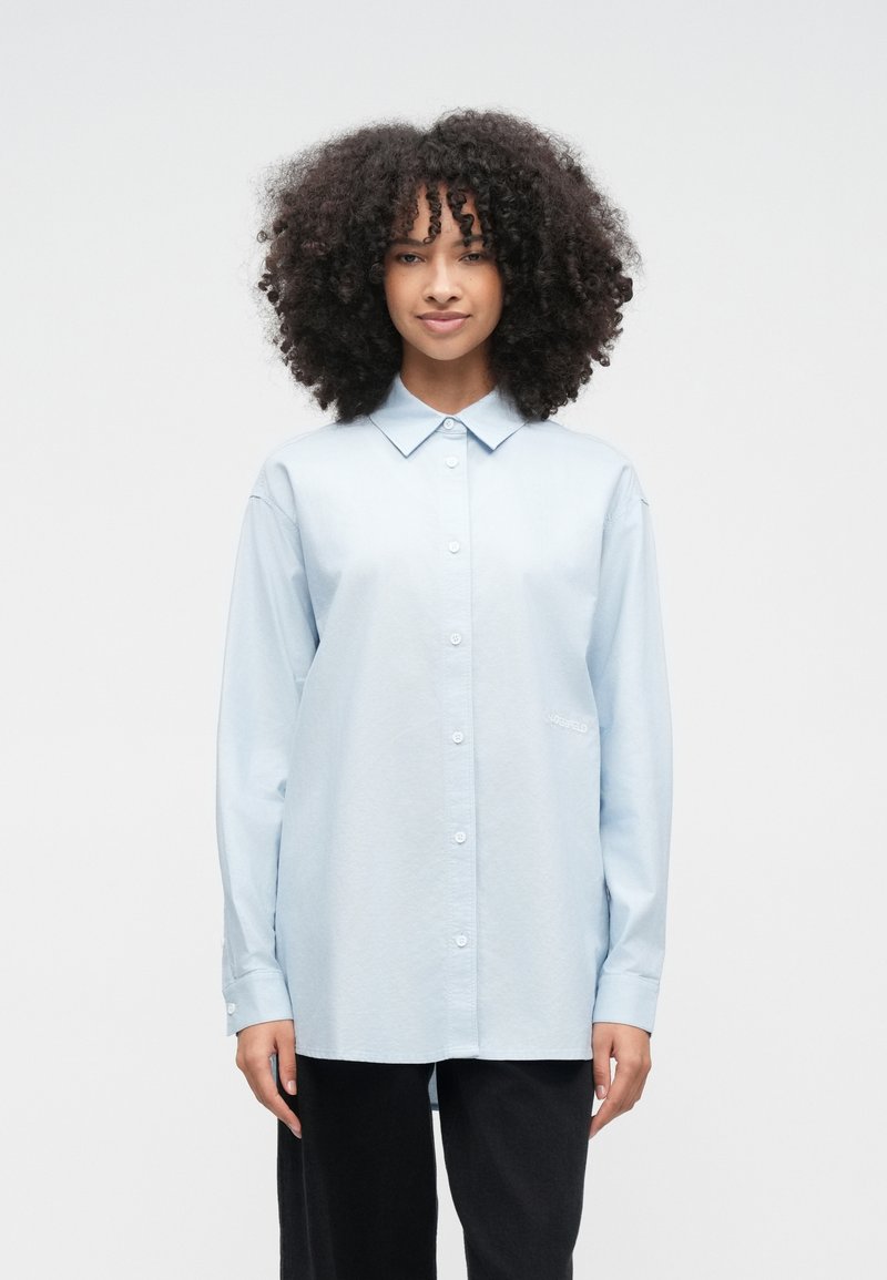 Kvinna med lockigt hår som bär en ljusblå skjorta med knappar och svarta byxor, stående mot en enkel vit bakgrund.