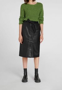 Jupe midi en cuir noir avec des boutons sur le devant et une taille nouée, associée à un pull en maille vert foncé et des bottes noires à talons épais.