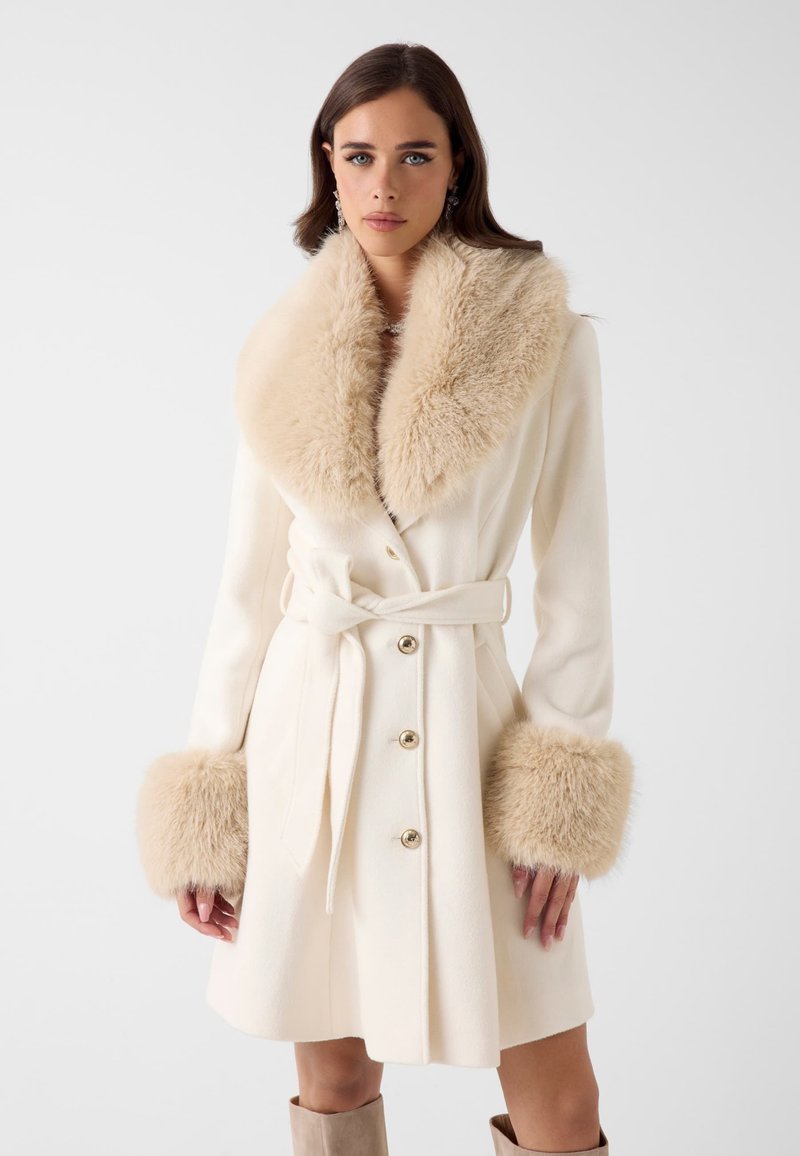Cappotto color crema con un grande colletto e polsini in eco pelliccia, vita sagomata e bottoni dorati, dal design strutturato ma fluido.