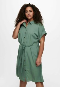 Zelena haljina s kratkim rukavima, kopčana sprijeda, s ovratnikom, remenom u struku i prorezom na dnu. Glatka tkanina i opušteni kroj.