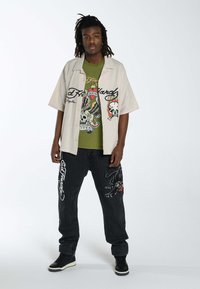 Camisa corta de color beige claro con letras negras, camiseta gráfica verde debajo, pantalones de mezclilla oscuros con diseños bordados y zapatillas negras.