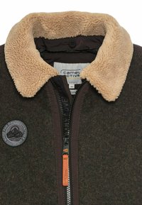 Brown fleece jacket with a beige shearling collar, zipper front, and a round patch featuring mountains. Soft texture and durable fabric.
