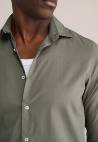 Olijfgroen overhemd met een zachte textuur, lange mouwen en een puntige kraag. Voorzien van knoopsluiting en een subtiele glans.