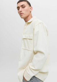 Jonge man in een crème corduroy shirt met borstzak, met zijn handen in zijn zakken, poserend tegen een effen lichte achtergrond.