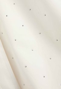 Witte stof met kleine, gelijkmatig verdeelde zilveren studs die een subtiel stippenpatroon vormen, met een close-up van textuur en detail.