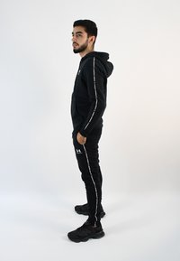 Conjunto deportivo negro que incluye una sudadera con capucha y pantalones ajustados. Ambos tienen acentos de logotipo blanco y texto a lo largo de los lados. Zapatillas deportivas negras.