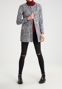 Cappotto in tweed grigio con linea dritta, collo rotondo e chiusura con bottoni, indossato sopra un dolcevita bordeaux e abbinato a leggings neri strappati e stivali.