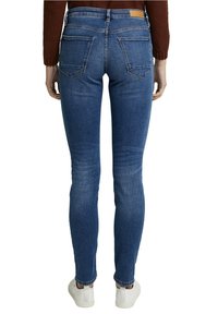 Jeans ajustados de mezclilla azul con una cintura alta, costuras visibles, dos bolsillos traseros y combinados con zapatillas blancas.