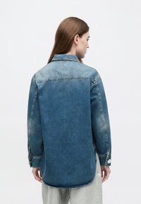 Γυναίκα με μακριά καστανά μαλλιά, που φοράει μπλε τζιν τζάκετ ombre και ανοιχτόχρωμο παντελόνι, γυρισμένη με την πλάτη προς ένα απλό φόντο.