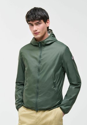 Giovane uomo che indossa una giacca a vento verde con cappuccio e zip, con le mani nelle tasche, in piedi davanti a uno sfondo chiaro uniforme.