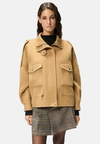 Veste en mélange de laine camel avec fermeture éclair et boutons-pression, grandes poches avant et épaulettes. Portée sur un pull noir et une jupe à carreaux.