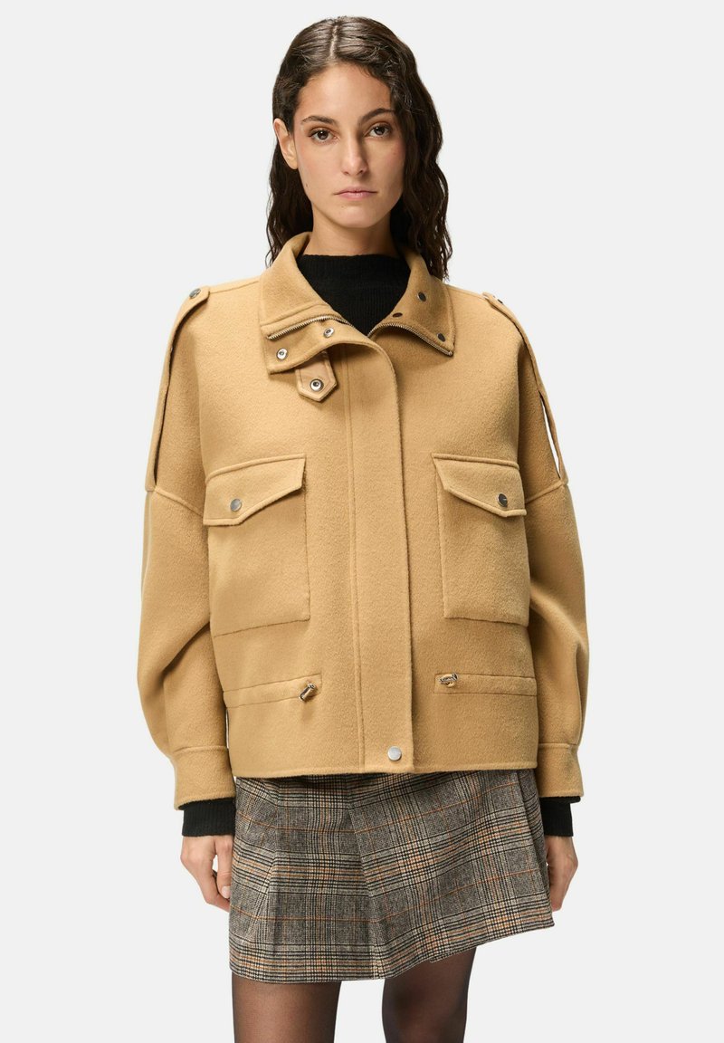 Veste en mélange de laine camel avec fermeture éclair et boutons-pression, grandes poches avant et épaulettes. Portée sur un pull noir et une jupe à carreaux.