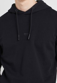 Sudadera negra hecha de tejido suave, con capucha ajustable y un pequeño logo en relieve en el centro. Textura suave, diseño minimalista.