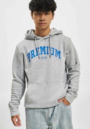 Ung mann med neseringer iført en grå hettegenser med blå "PREMIUM" tekst, lyse denimjeans, stående med en hånd i lommen.