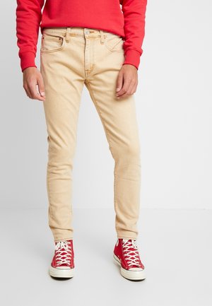 Beige slim-fit farkut, joissa on viisi taskua, vyölenkit ja vaaleat ompelusyksityiskohdat, yhdistettyinä punaisiin korkeisiin tennareihin.
