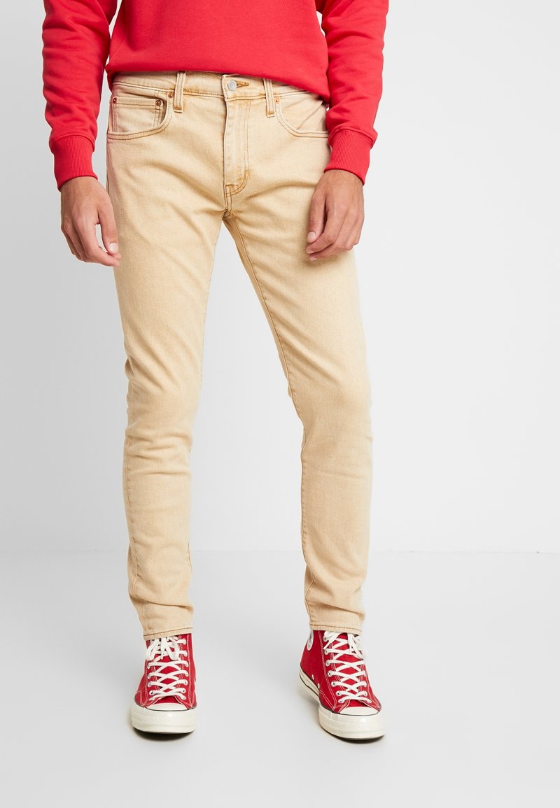 Beige slim-fit jeans with five pockets, belt loops, and light stitching details, paired with red high-top sneakers.
