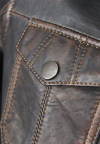 Nahaufnahme einer braunen Lederjacke mit einem silbernen Druckknopf und kontrastierendem gelbem Stich auf einer strukturierten Oberfläche.