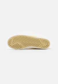 Semelle en caoutchouc de sneaker avec un motif zigzag beige, ornée d'un contour blanc. La texture indique une bonne adhérence et durabilité, conçue pour la traction.