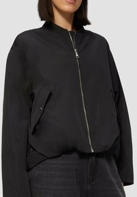 Veste bomber noire en tissu lisse, dotée d'une fermeture éclair sur le devant, de poches latérales et d'un col côtelé. Design simple et moderne.