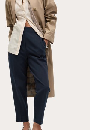 Persoon draagt een beige trenchcoat, een gestreept overhemd waarvan de voorkant gedeeltelijk uit de broek steekt, en een hooggewaste marineblauwe broek met handen in de zakken.