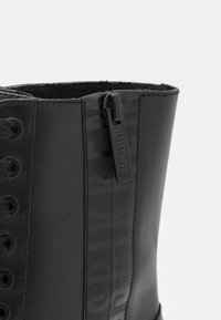 Gros plan d'une botte en cuir noir avec fermeture éclair latérale et lacets, présentant le nom de la marque embossé sur le zip et la bande.