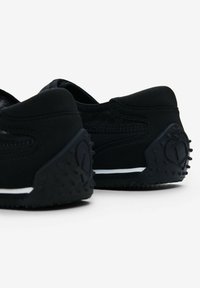 Black athletic shoes with a textured fabric upper, rear rubber heel with protrusions, subtle white stripe on the sole, and logo detail.