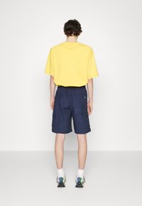 Žluté oversize tričko v kombinaci s námořnickými cargo šortkami. Boty mají vícebarevný design. Hladký bílý pozadí.