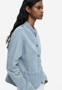 Svijetloplava prugasta jakna s ovratnikom, kop čanjem na gumbe i prednjim džepovima. Izrađena od teksturiranog materijala s kontrastnim vertikalnim prugama.