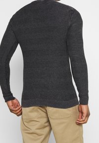 Czarny sweter dzianinowy z teksturowanym wzorem, z długimi rękawami oraz ściągaczami na dole i przy mankietach, zestawiony z beżowymi spodniami.