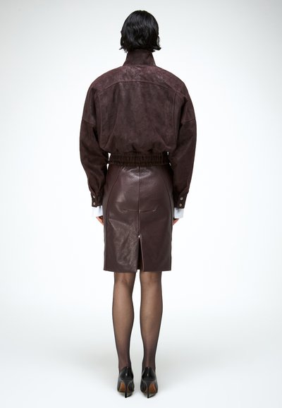 Osoba viđena sa stražnje strane u tamnobraon antilop jakni i odgovarajućoj kožnoj suknji s prorezom u sredini, prozirnim crnim čarapama i crnim potpeticama.