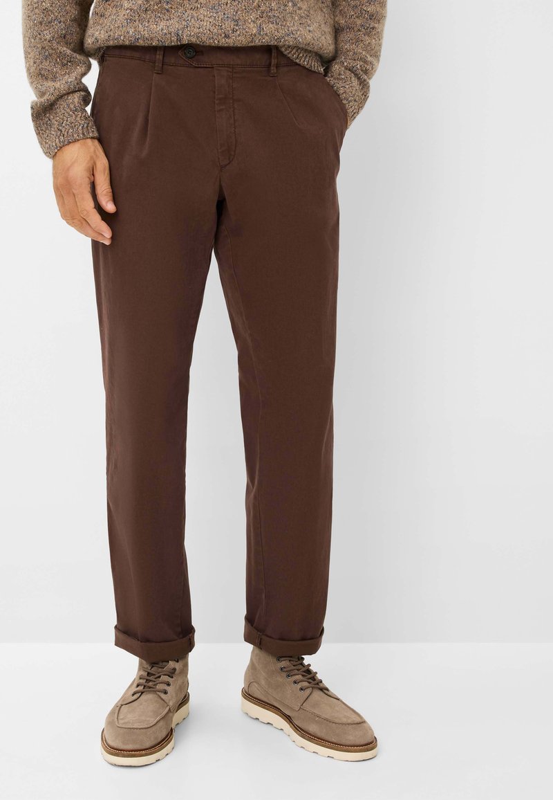 Braune Baumwollhose mit Falten, gesäumten Bündchen und Knopfverschluss. Kombiniert mit hellbeigen Wildlederschuhen mit Schnürsenkeln.