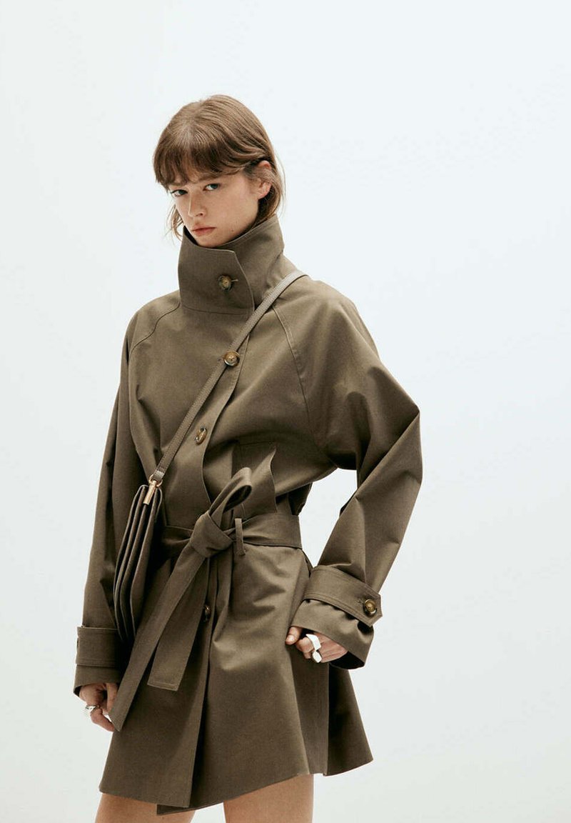 Femme portant un manteau ceinturé vert olive à col montant et un sac bandoulière, debout devant un fond clair uni.