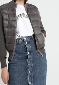 Mujer con una chaqueta acolchada gris sobre una camiseta gráfica blanca y una falda de mezclilla de botón delantero y tiro alto.