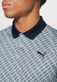 Ljusblå pikétröja med lövmönster, marinblå krage och tre vita knappar. Har en svart Puma-logga på vänster sida.