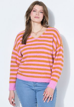 Kvinde med langt brunt hår iført en lyserød og orange stribet sweater og lyseblå jeans, stående foran en hvid baggrund.