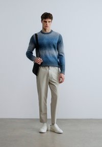 Sinisen liukuvärjätty neule, jossa resorinen kaula-aukko, beiget nyörillä säädettävät housut ja valkoiset tennarit. Malli pitää kädessään mustaa olkalaukkua.