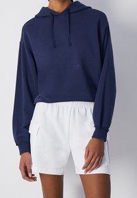 Námořnická zkrácená mikina s kapucí a dlouhými rukávy, doplněná bílými šortkami s elastickým pasem, které mají boční kapsy a jemnou texturu.