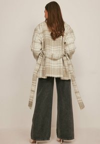 Grijs-witte geruite oversized wikkelcardigan met brede mouwen, een strikceintuur en een gestructureerde breiwijze. Gecombineerd met donkere jeans met wijde pijpen.