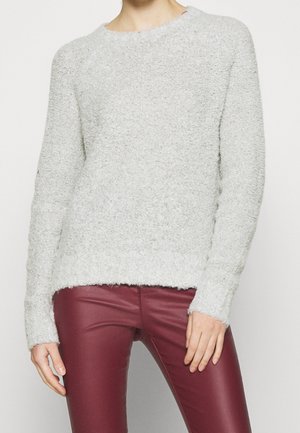 Persoon die een lichtgrijze, getextureerde trui met lange mouwen en nauwsluitende bordeauxrode leren broek draagt, staand tegen een effen achtergrond.