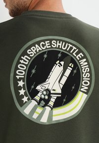 Felpa verde oliva con un grafico circolare di un shuttle spaziale, la scritta "100° MISSIONE DELLO SPACE SHUTTLE" e accenti di stelle bianchi.