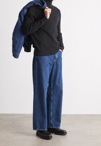 Schwarzer Pullover mit Strukturstrick, lockere blaue Jeans mit weitem Bein und schwarze robuste Stiefel. Blaue Jeansjacke lässig  über eine Schulter gelegt.