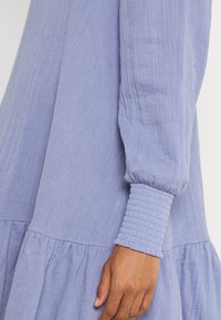 Vaaleanlila mekko, jossa on teksturoitu kangas, kerätty vyötärö ja puffihihat, viimeistelty ribatulla yksityiskohdalla hihojen suissa.