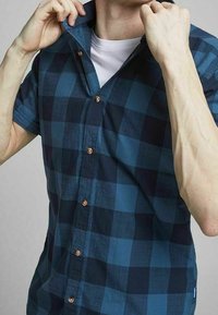 Man i en blå och svart rutig kortärmad skjorta över en vit t-shirt, som justerar kragen med båda händerna.