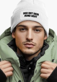 White knit beanie with a black text patch reading "DER MIT DEM HUND GEHT"; worn under an olive green, zipped puffer jacket layered over a black top.