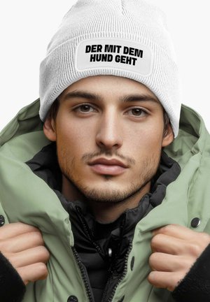 White knit beanie with a black text patch reading "DER MIT DEM HUND GEHT"; worn under an olive green, zipped puffer jacket layered over a black top.