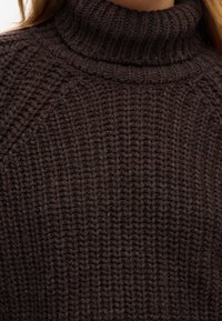 Gros plan sur un pull col roulé en maille épaisse de couleur brun foncé porté par une personne à la peau claire et aux cheveux longs.