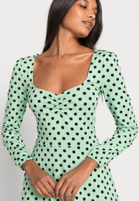 Vestido verde menta com mangas longas, apresentando um decote quadrado e bolinhas pretas. O tecido tem um leve brilho e um detalhe franzido na zona do peito.