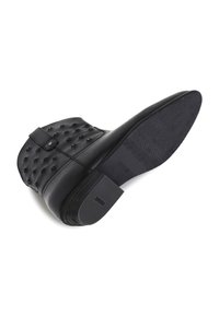 Bottine noire en cuir avec un design texturé et clouté. Elle possède une semelle plate, une sangle décorative et un talon carré.