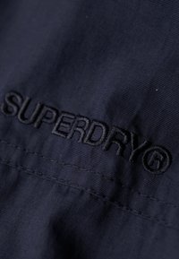 Marinblåt stof med en glat tekstur og broderet "SUPERDRY" logo. Designet inkluderer syede detaljer og et rent, minimalistisk look.